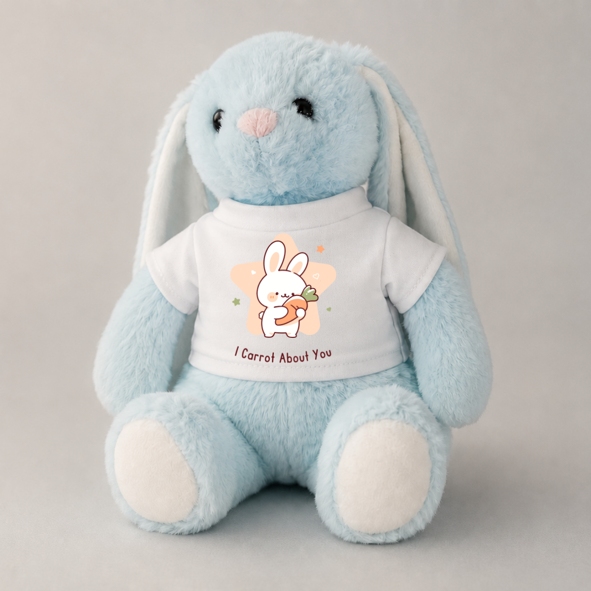 A light blue plush bunny toy with long floppy ears, a pink nose, and white paws, wearing a white T-shirt printed with a cartoon bunny holding a carrot and the text 'I Carrot About You'.