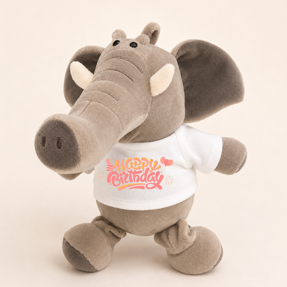A plush elephant toy in warm gray-brown fabric wearing a white T-shirt with a pink and orange 'Happy Birthday' graphic, photographed against a plain light beige background.