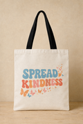 An off-white canvas tote bag with black shoulder straps, featuring the phrase 'SPREAD KINDNESS' in blue and orange lettering with small colorful butterflies, set against a warm beige background.