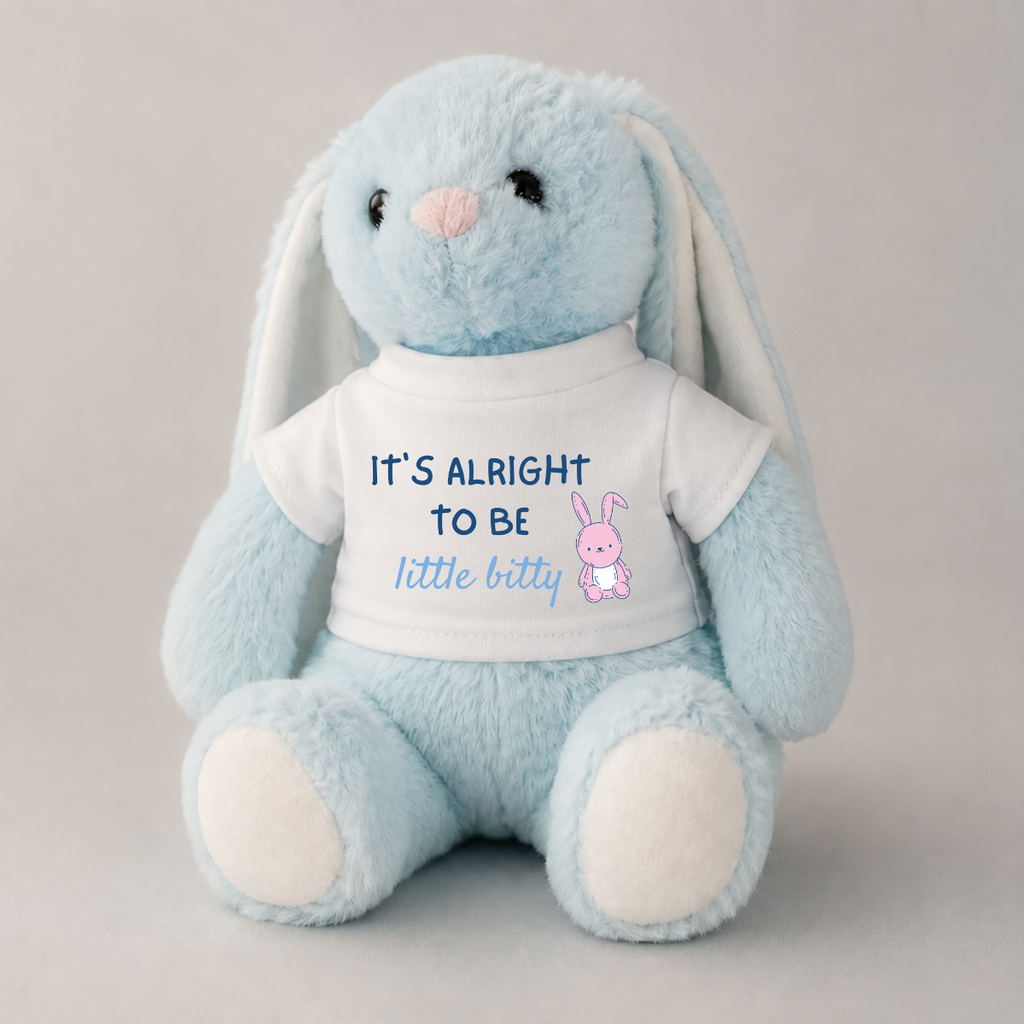 A light blue plush bunny toy with long floppy ears, a pink nose, and a white T-shirt printed with the text 'IT'S ALRIGHT TO BE little bitty' and a small pink bunny graphic, photographed against a neutral light gray background.