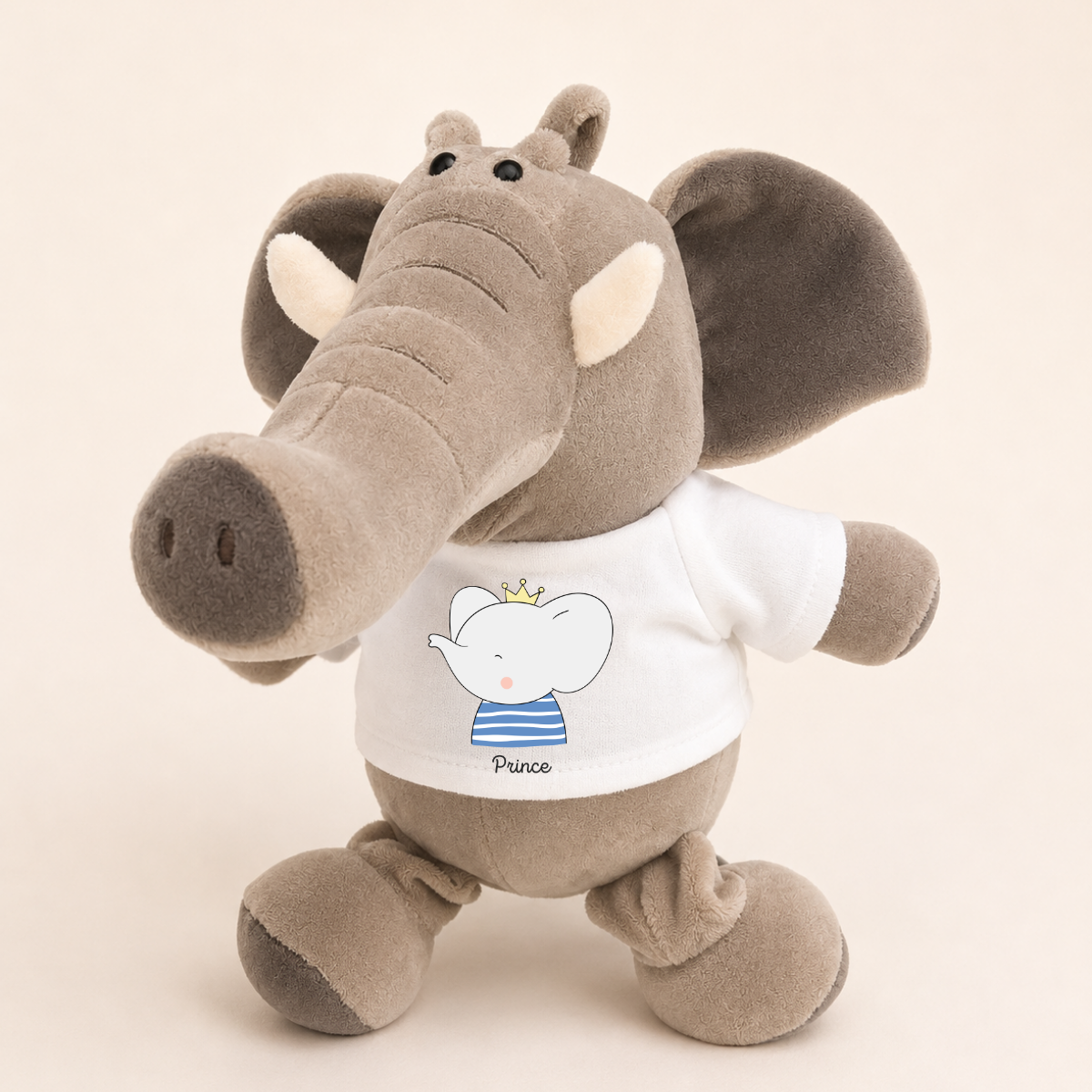 A plush elephant toy in warm gray-brown fabric wearing a white T-shirt with a cartoon baby elephant and the word 'Prince' on the front, photographed against a plain light beige background.