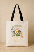 An off-white canvas tote bag with black shoulder straps, featuring a printed graphic of wildflowers in a pocket with the text 'WILDFLOWER SOUL' on a beige background.