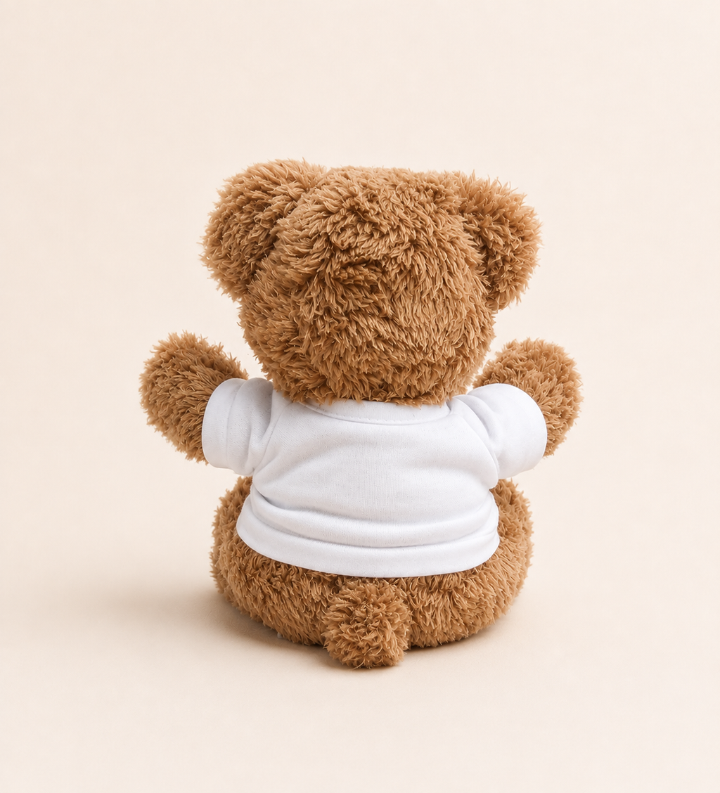 A brown plush teddy bear viewed from the back, wearing a plain white short-sleeve shirt, centered on a warm cream background.