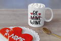 White mug with 'Be Mine Wine' text, heart symbols, and a cookie shaped like a heart with text on a wooden surface.