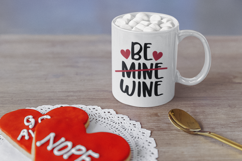 White mug with 'Be Mine Wine' text, heart symbols, and a cookie shaped like a heart with text on a wooden surface.
