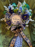 Decorative wreath with flowers and a 'Welcome' sign on a green door.