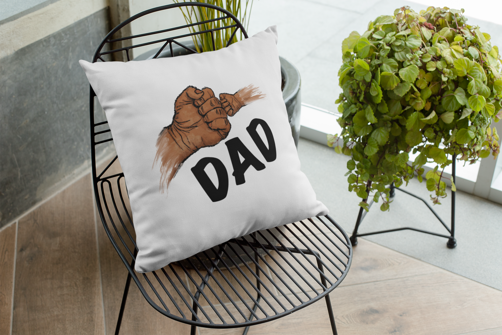 Decorative pillow with 'DAD' and hand graphic on a black wire chair with plants in the background.