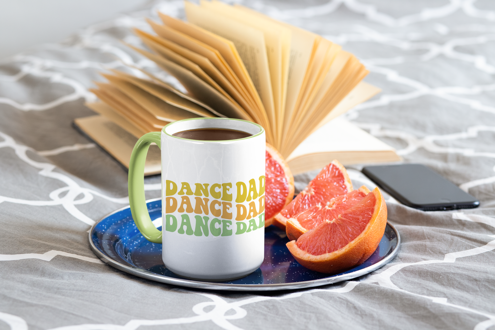 White mug with 'DANCE DAD' text on a saucer with fruit and a phone, on a textured surface.