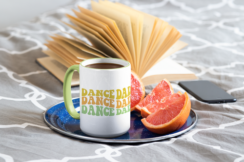 White mug with 'DANCE DAD' text on a saucer with fruit and a phone, on a textured surface.