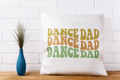 White pillow with 'DANCE DAD' text on a wooden surface with a white brick wall background