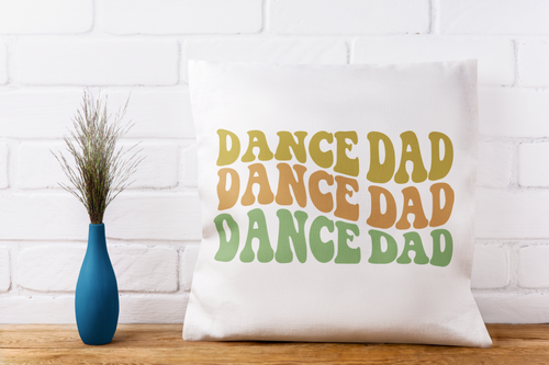 White pillow with 'DANCE DAD' text on a wooden surface with a white brick wall background