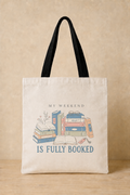 A cream canvas tote bag with black shoulder straps, featuring a cute illustration of stacked books and the text 'MY WEEKEND IS FULLY BOOKED' in blue and black, set against a warm beige background.
