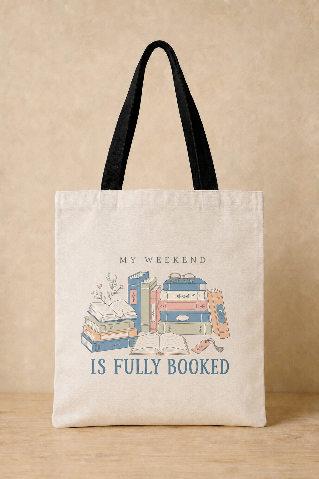 A cream canvas tote bag with black shoulder straps, featuring a cute illustration of stacked books and the text 'MY WEEKEND IS FULLY BOOKED' in blue and black, set against a warm beige background.