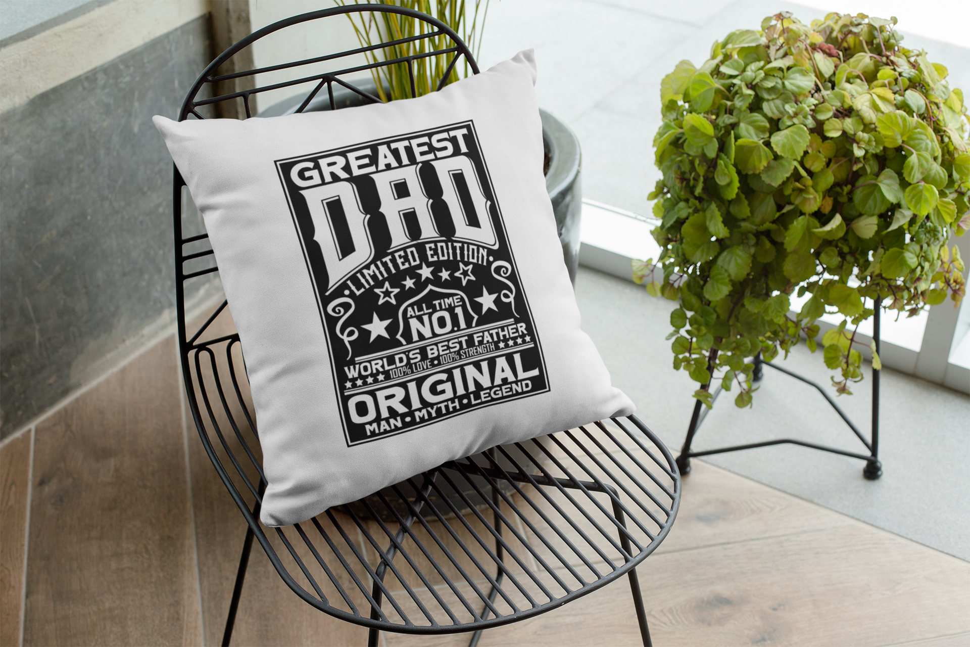 Pillow with 'Greatest Dad' text on a black wire chair with plants in the background