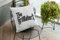 White pillow with 'The Grillfather' text on a black wire chair with plants in the background.