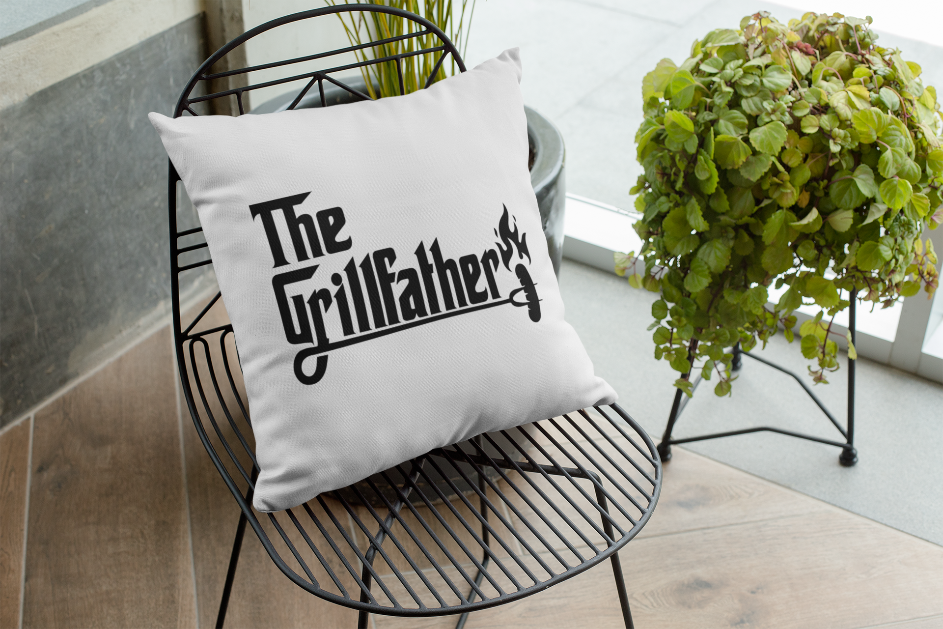 White pillow with 'The Grillfather' text on a black wire chair with plants in the background.