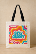An off-white canvas tote bag with black shoulder straps, featuring a colorful retro psychedelic wavy pattern in pink, orange, yellow, and blue with the text 'STAY GROOVY' in light blue. The bag is displayed standing upright on a light wood surface against a textured beige wall.