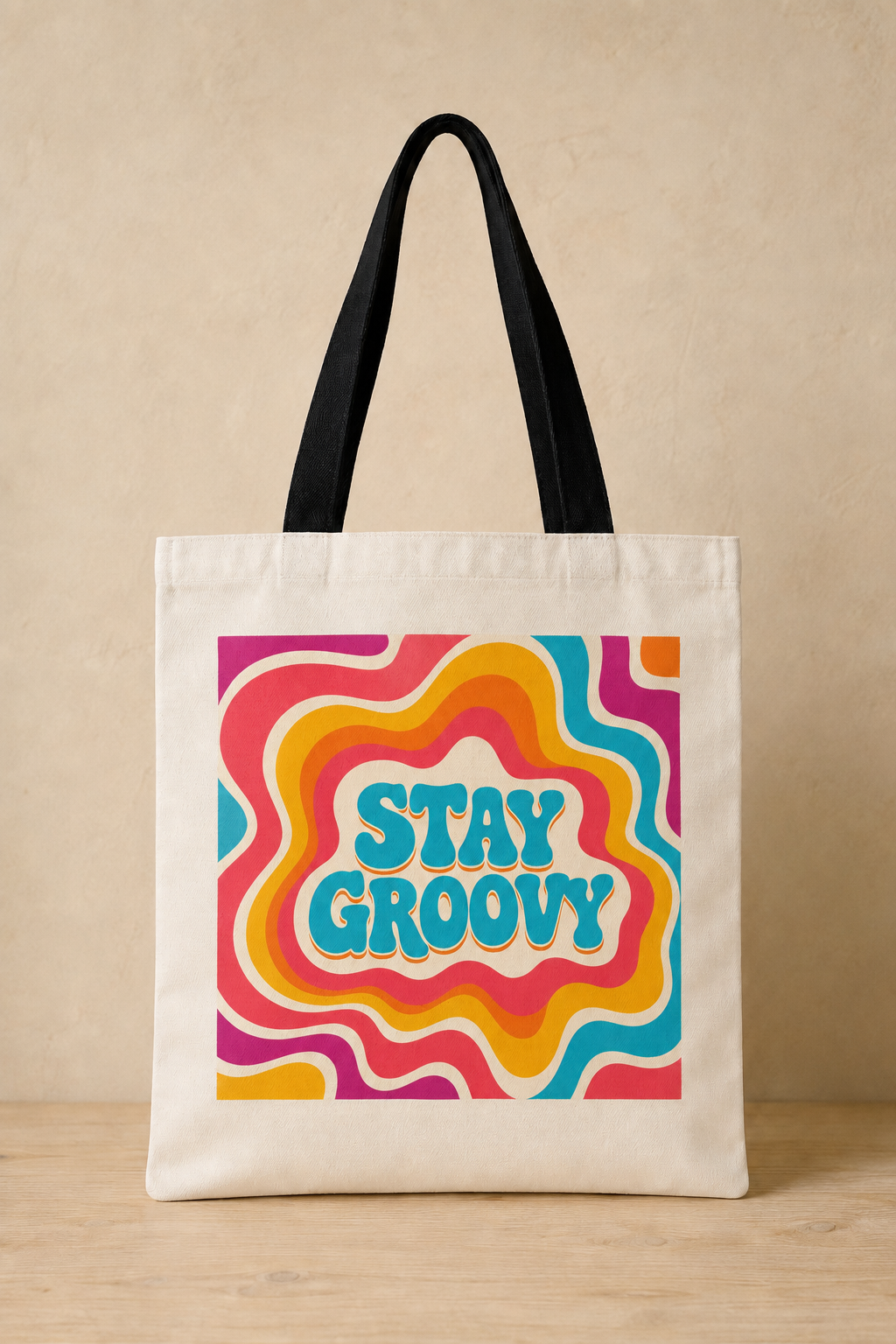 An off-white canvas tote bag with black shoulder straps, featuring a colorful retro psychedelic wavy pattern in pink, orange, yellow, and blue with the text 'STAY GROOVY' in light blue. The bag is displayed standing upright on a light wood surface against a textured beige wall.