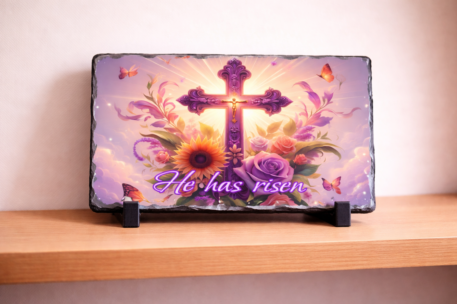 Decorative slate with a cross, flowers, and 'He has risen' text on a wooden shelf.