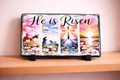 Decorative slate with 'He is Risen' text and Easter-themed illustrations on a wooden shelf.