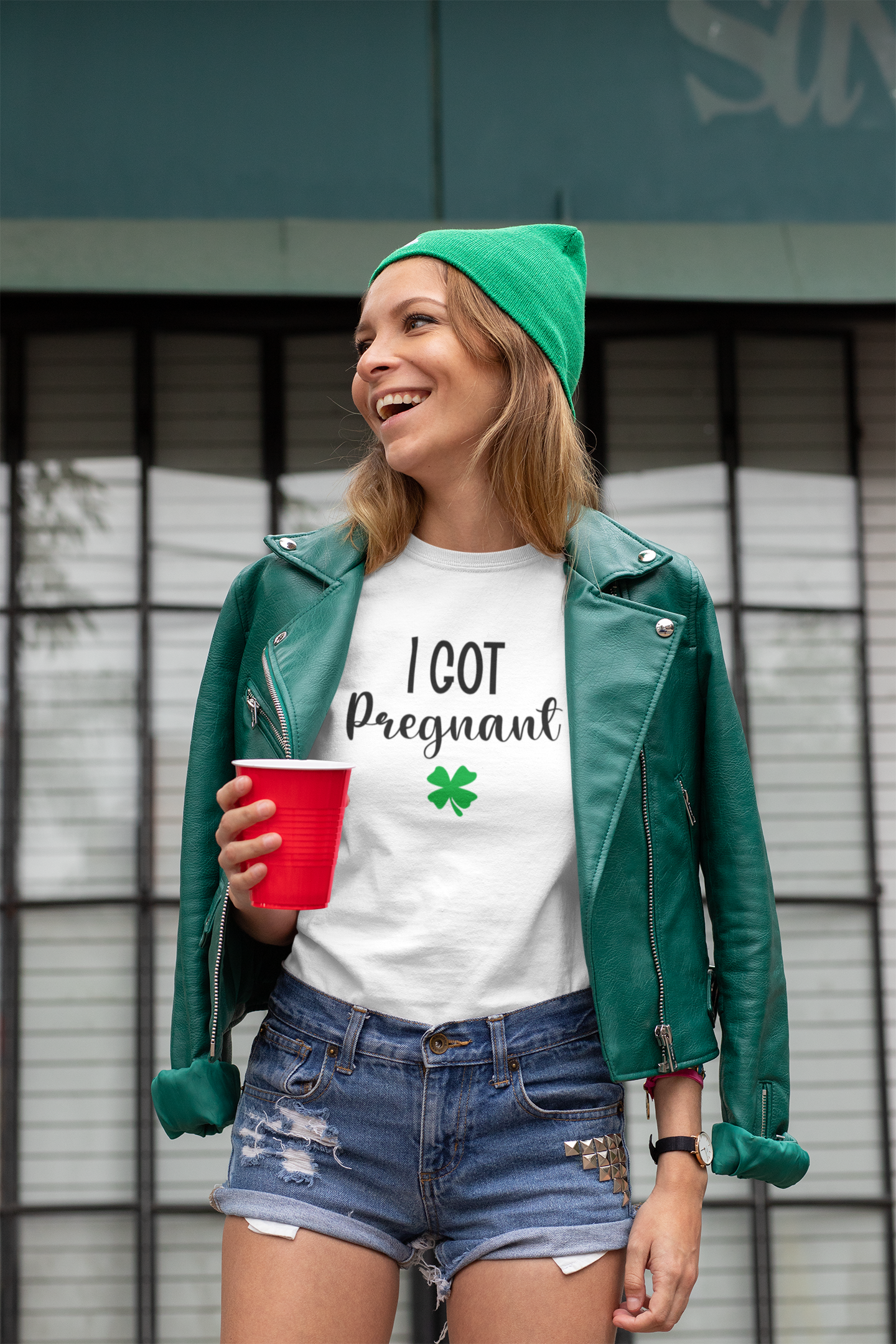 Woman wearing a green beanie and leather jacket, holding a red cup, with text on her shirt.
