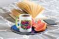 Mug with floral design and 'Mom' text on a saucer with fruit and phone, on a textured surface.