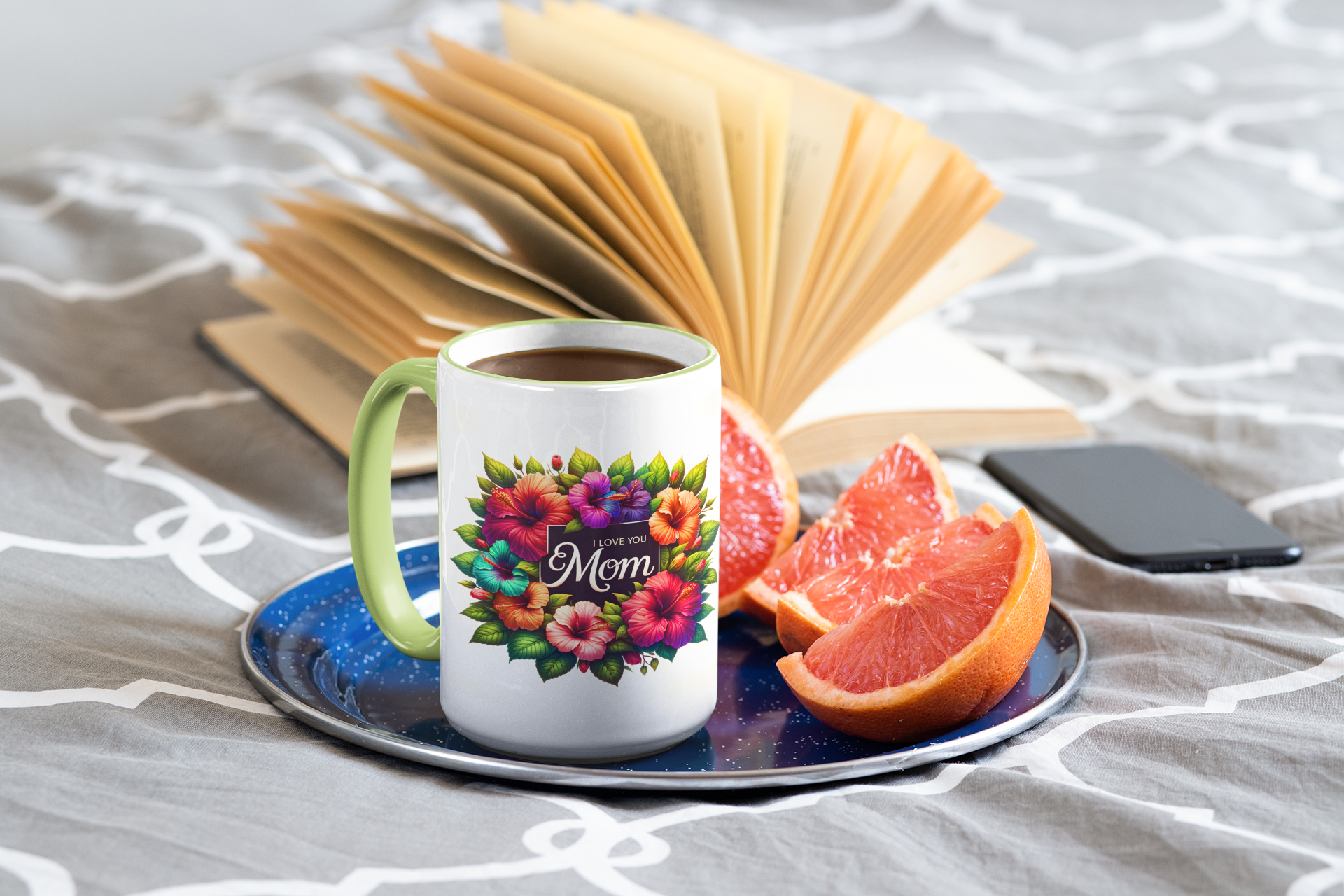 Mug with floral design and 'Mom' text on a saucer with fruit and phone, on a textured surface.
