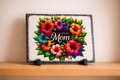 Decorative plaque with colorful flowers and 'I Love You Mom' text on a wooden surface.