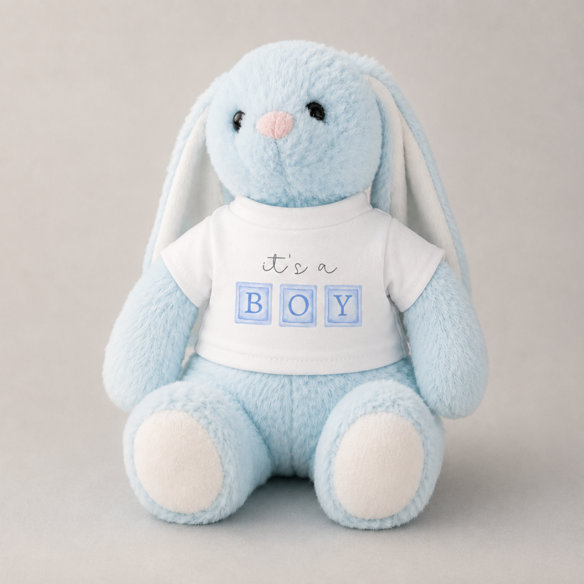 A light blue plush bunny toy wearing a white T-shirt that reads 'it's a BOY' with the letters in blue blocks, photographed against a soft light gray background.
