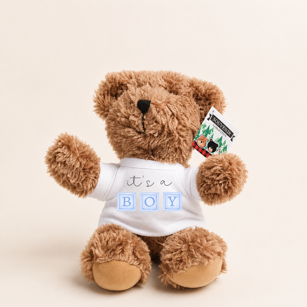 A brown plush teddy bear wearing a white t-shirt that reads 'it's a BOY' in blue block letters, sitting against a light beige background. A small 'MERRY CHRIST' gift tag is attached to its ear.