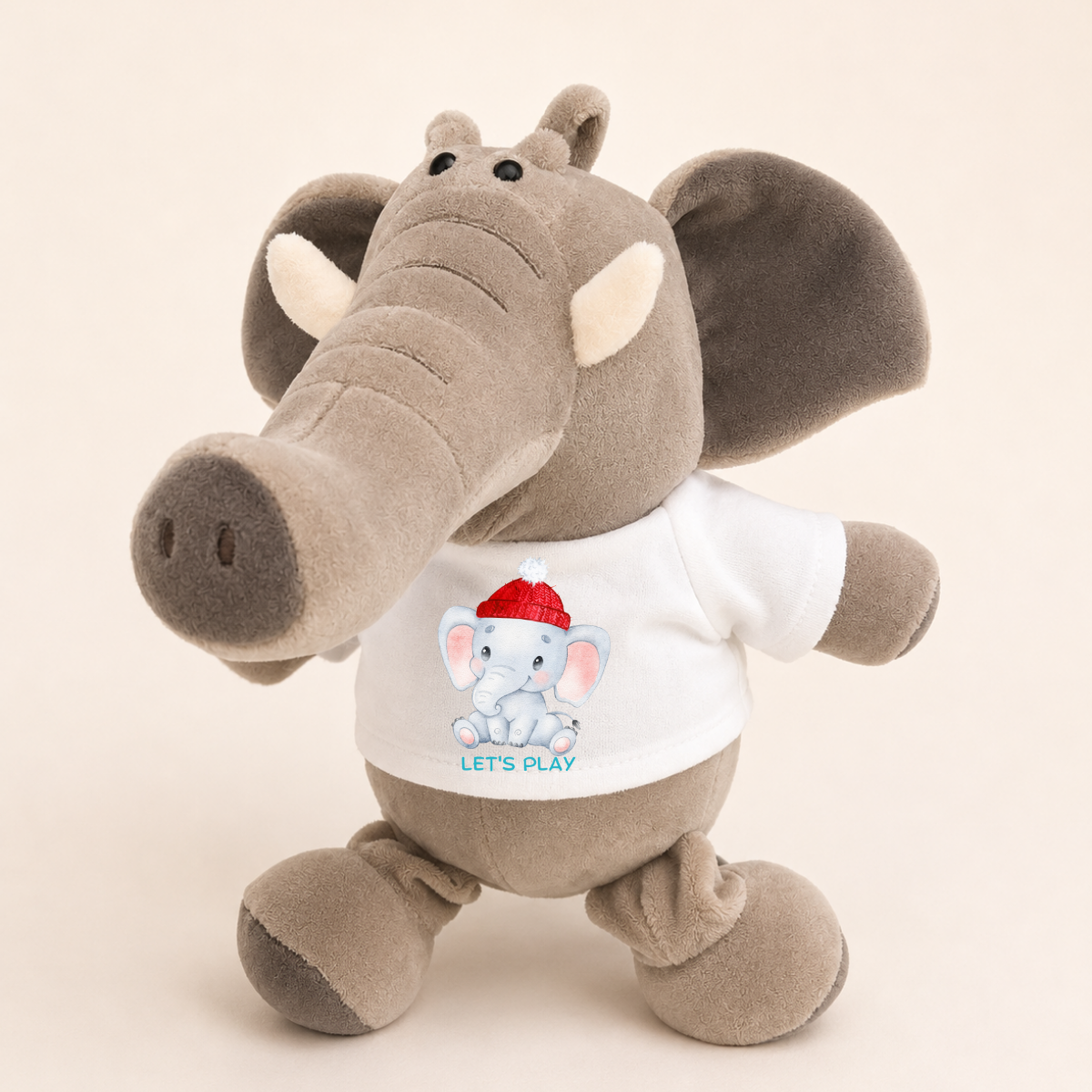 A plush toy elephant in gray fabric wearing a white T-shirt with a cartoon baby elephant graphic and the words 'LET'S PLAY' on the front, photographed against a plain light beige background.