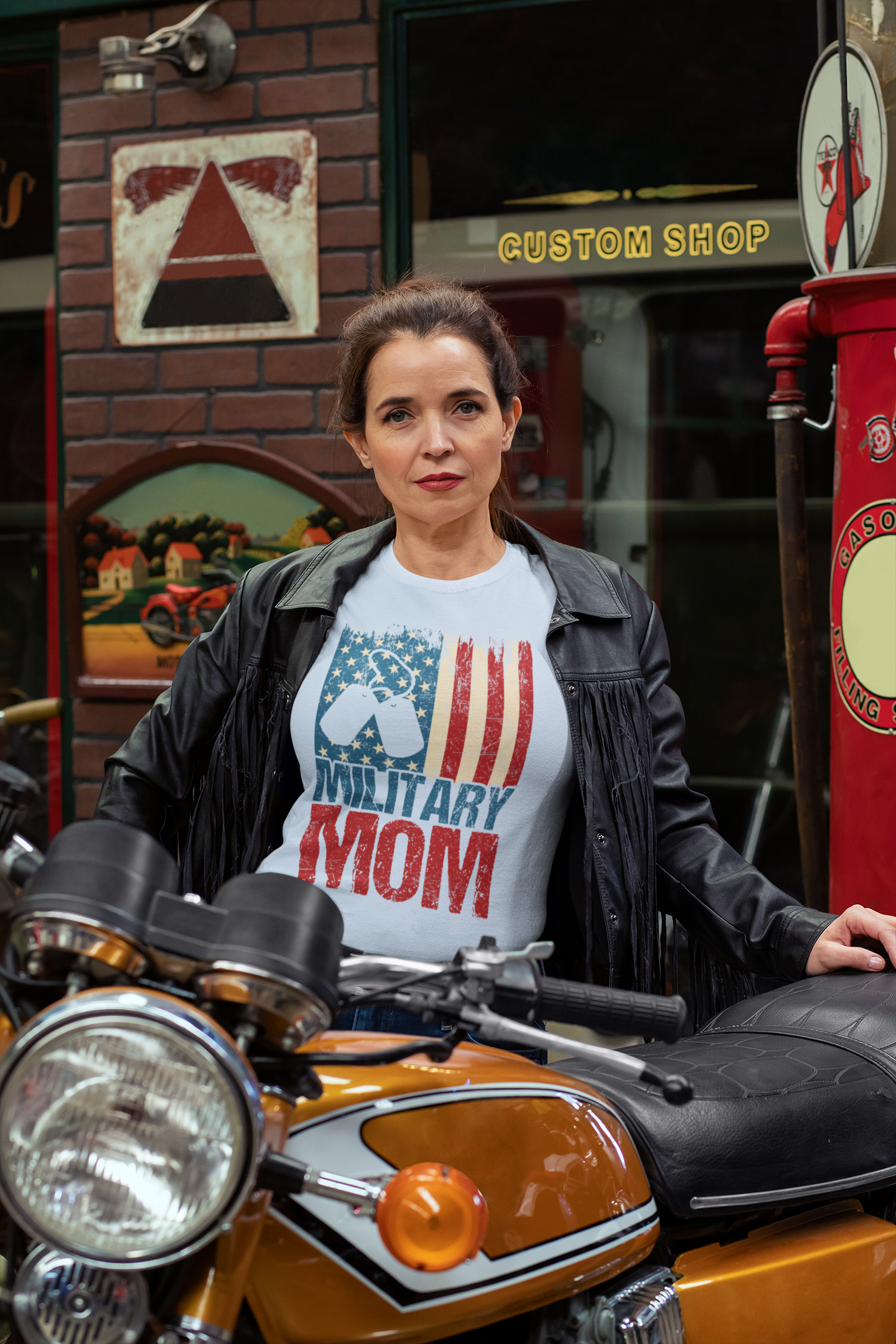 Woman wearing a 'Military Mom' t-shirt sitting on a motorcycle in front of a building labeled 'Custom Shop'.