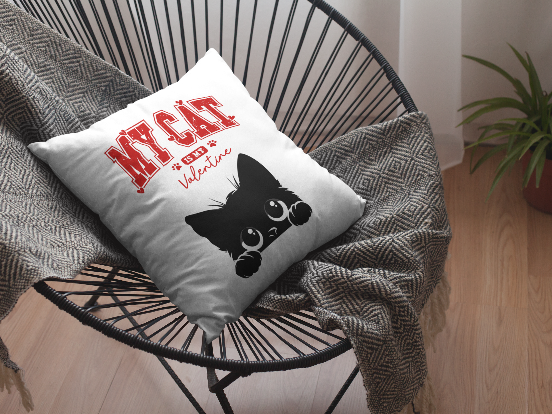 Decorative pillow with a black cat illustration and text on a chair with a blanket and plant in the background