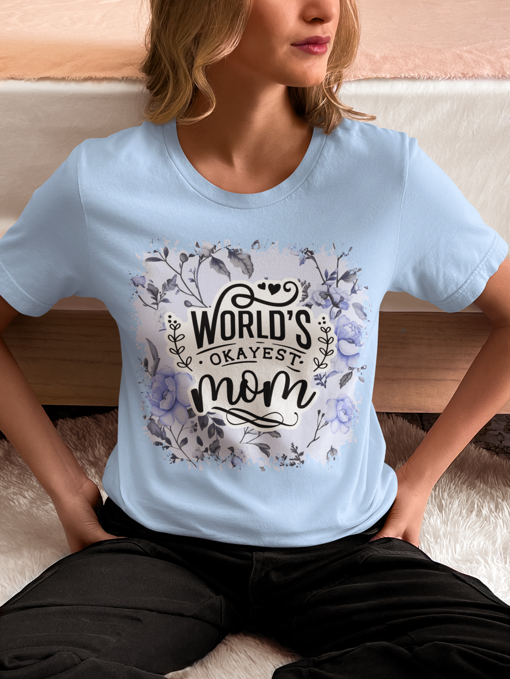 Person wearing a light blue t-shirt with 'World's Okayest Mom' text and floral design.