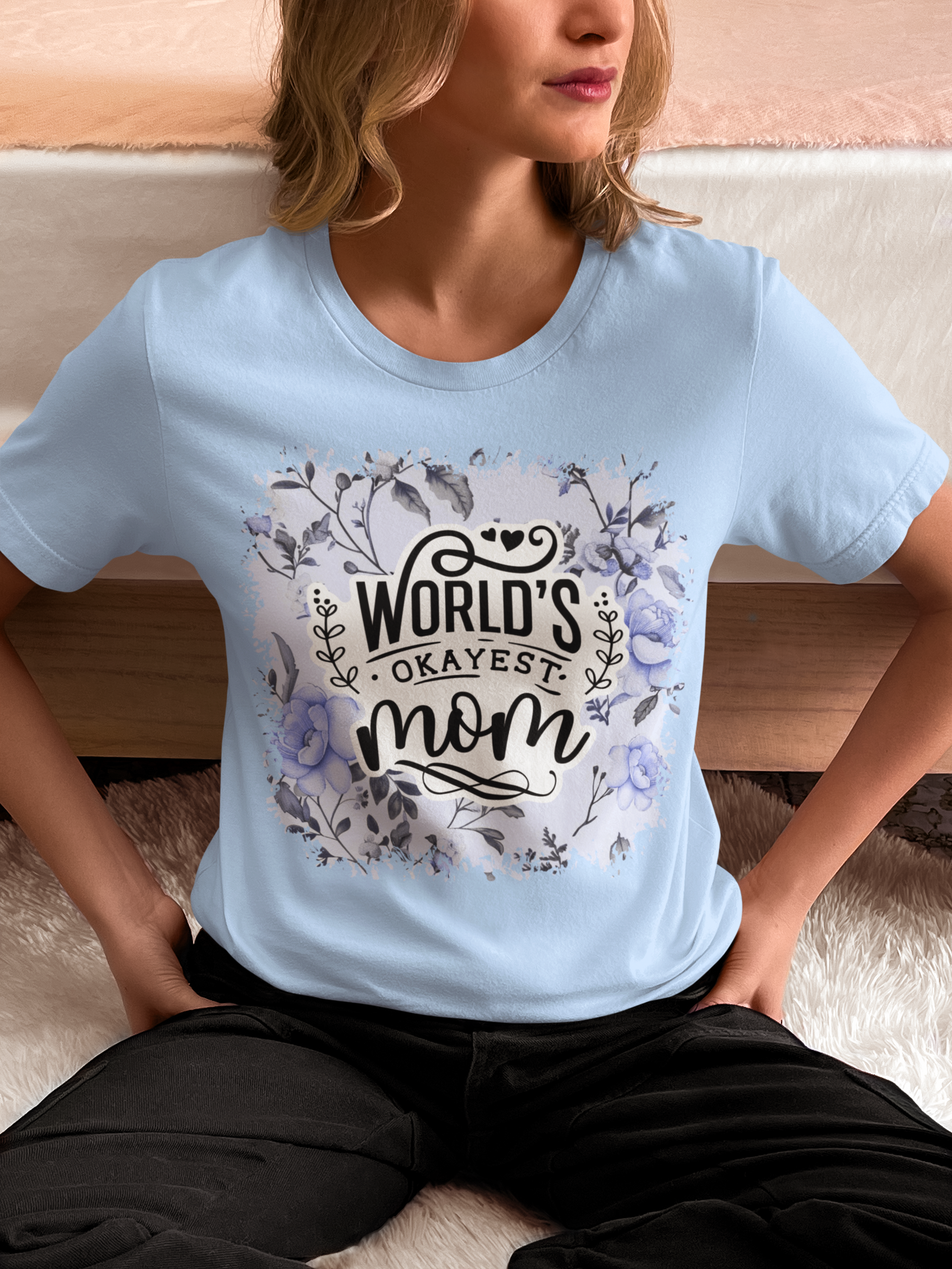 Person wearing a light blue t-shirt with 'World's Okayest Mom' text and floral design.