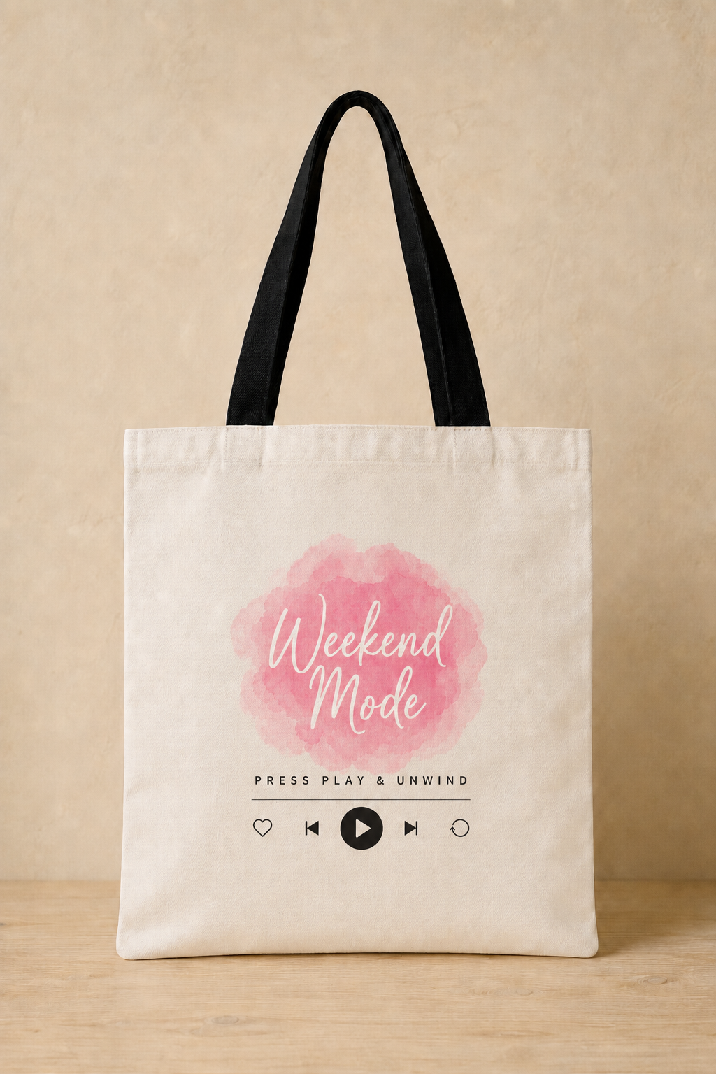 A cream canvas tote bag with black shoulder straps, featuring a pink watercolor circle and the words 'Weekend Mode' with music player icons on the front, set against a warm beige background.