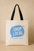 An off-white canvas tote bag with black shoulder straps, featuring a blue speech bubble graphic with the words 'SHOP LOCAL' and smaller 'SUPPORT LOCAL' text below, set against a warm beige background.