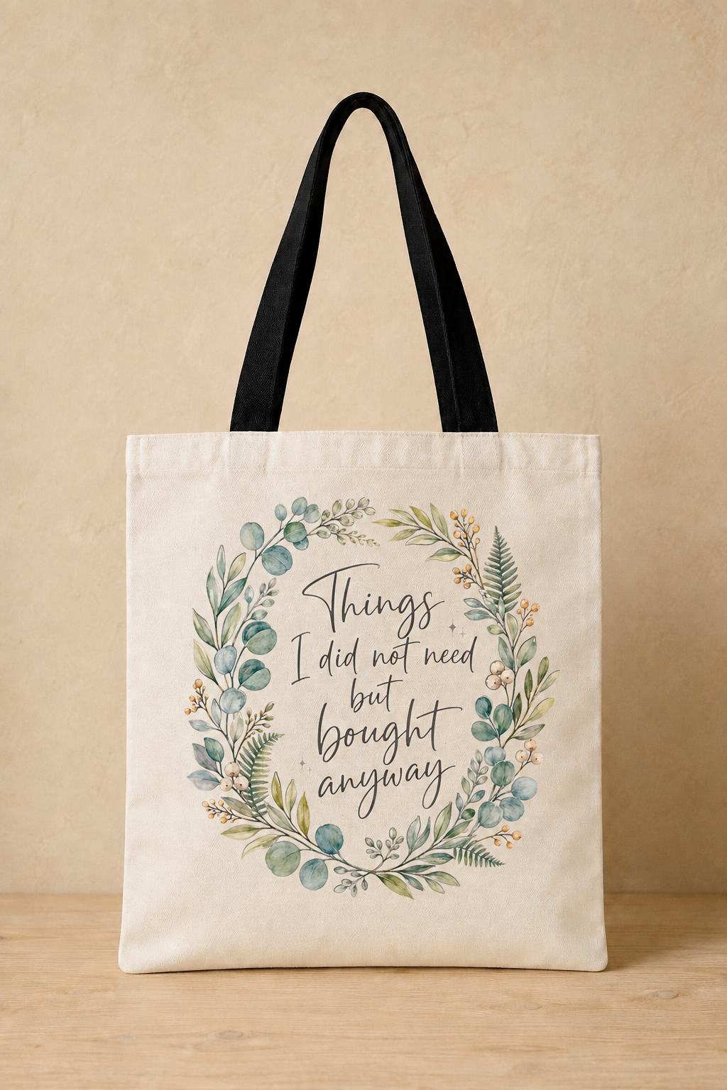 An off-white canvas tote bag with black shoulder straps, featuring a watercolor floral wreath and the quote 'Things I did not need but bought anyway' printed on the front, set against a warm beige background.