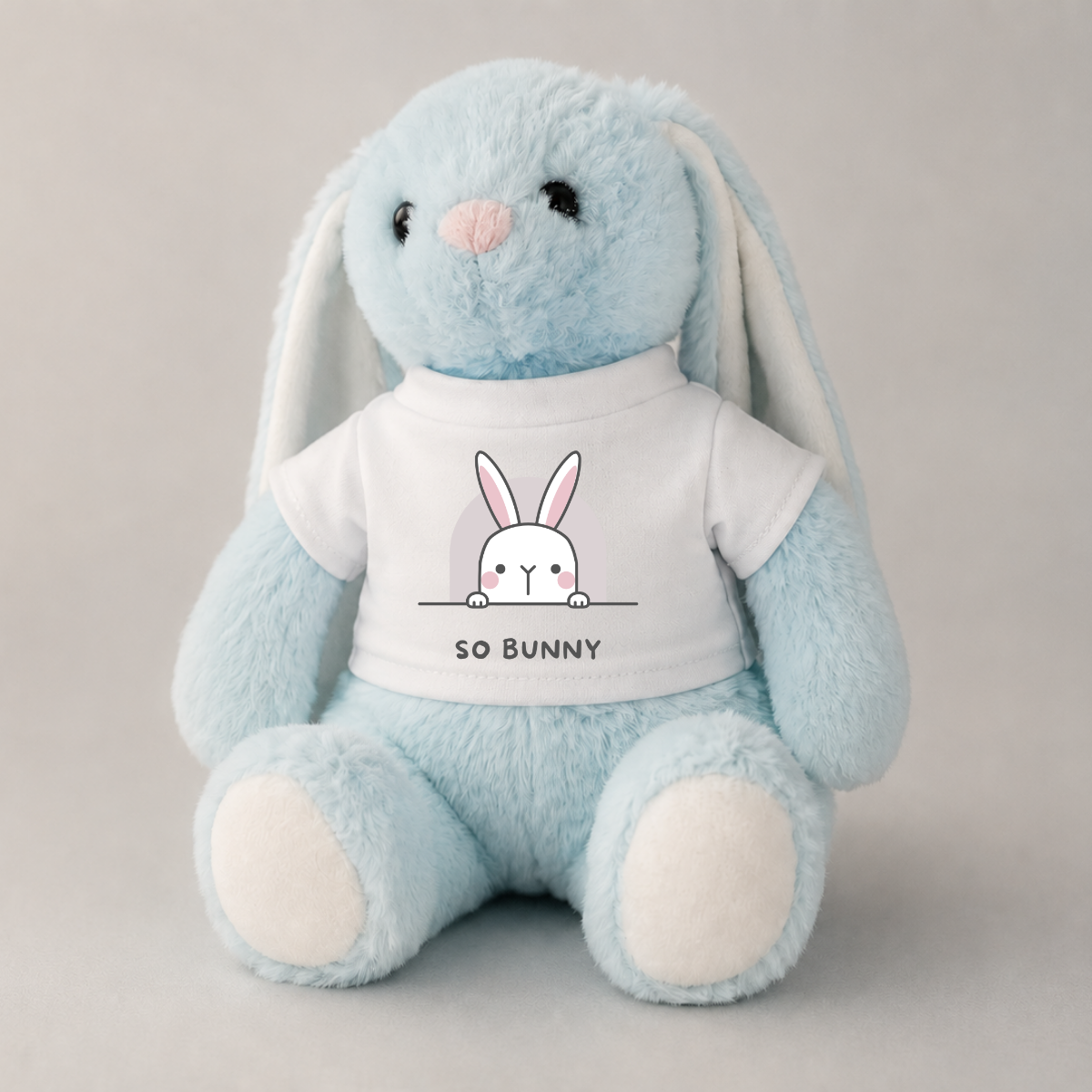 A light blue plush bunny toy with long floppy ears, a pink nose, and white paw pads, wearing a white T-shirt printed with a cartoon bunny peeking over a line and the text 'SO BUNNY'.