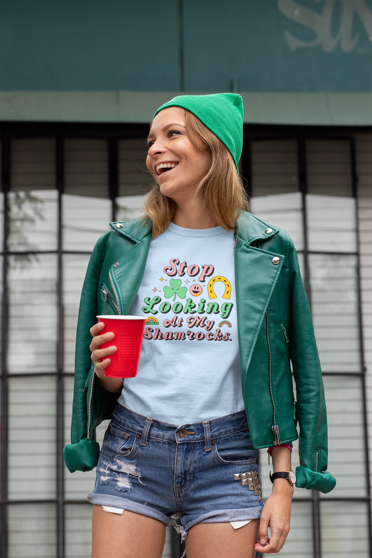 Person wearing a green beanie and leather jacket, holding a red cup, with a blue t-shirt in front of a building.