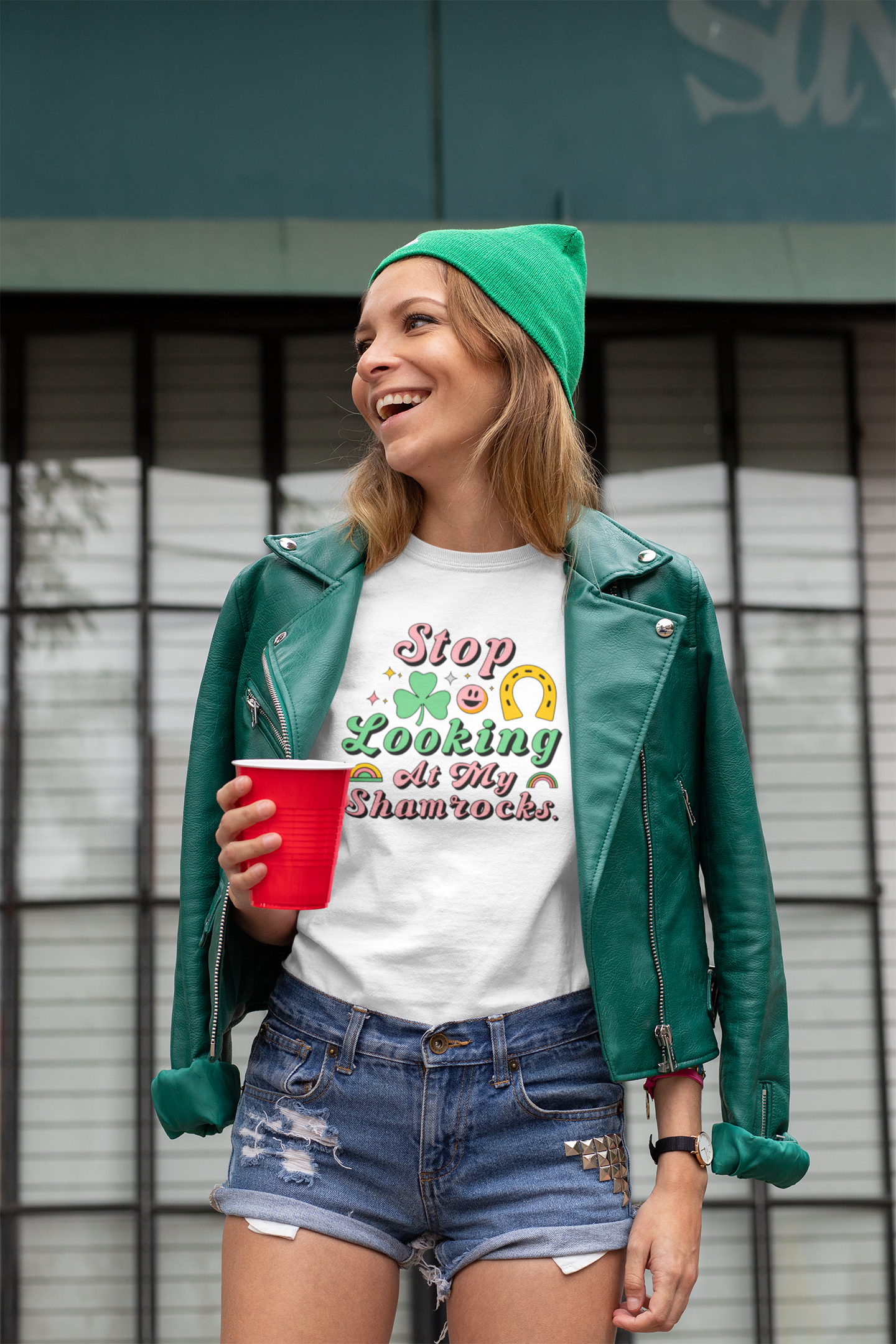 Person wearing a green beanie and leather jacket, holding a red cup, with a white t-shirt featuring colorful text.